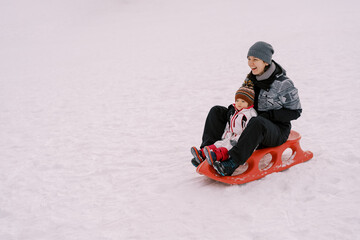 Fototapeta na wymiar Laughing mother with a little girl ride a sleigh on a snowy hill