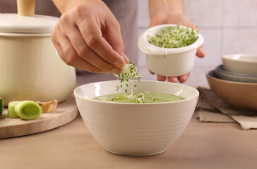 Woman adding fresh microgreens into bowl with tasty leek soup at wooden table, closeup