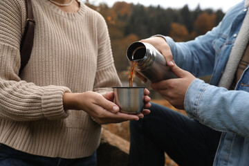 Naklejka premium Boyfriend pouring hot drink from metallic thermos into cup lid for his girlfriend outdoors, closeup