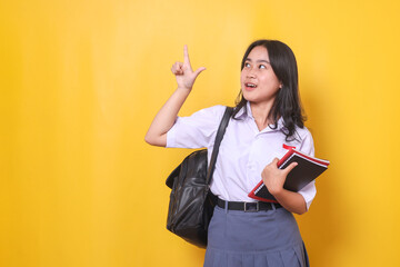 Young Asian student girl in high school uniform, holding book and pointing above to the free space for educational ads