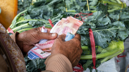 an old woman's hand holding a rupiah note with a denomination of Rp.100,000 against a background of green broccoli vegetables - obrazy, fototapety, plakaty