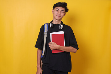 Portrait of handsome Asian student carrying backpack and holding books on yellow background