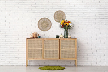 Wooden cabinet with bouquet of autumn flowers and pumpkins near white brick wall
