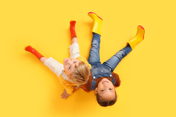 Little children in rubber boots sitting on yellow background, top view