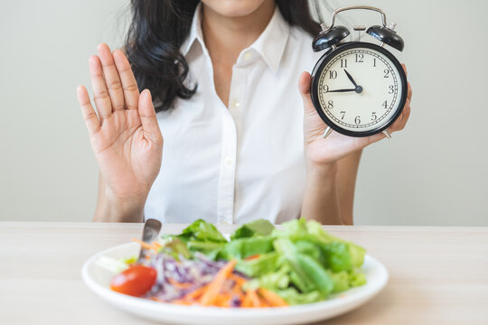 Intermittent fasting with clock, asian young woman dieting hand refuse waiting time to eat low carb food, green vegetable salad on plate. Eat food healthy first meal on brunch, lunch on table at home.