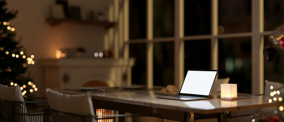 A laptop mockup on a dining table in a living room with a fireplace and a Christmas tree at night.