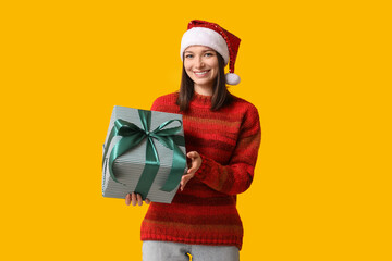 Beautiful young woman in Santa hat with gift box on yellow background