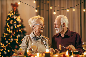 A festive senior couple eating at christmas table and celebrating christmas and new year's eve at home. - obrazy, fototapety, plakaty