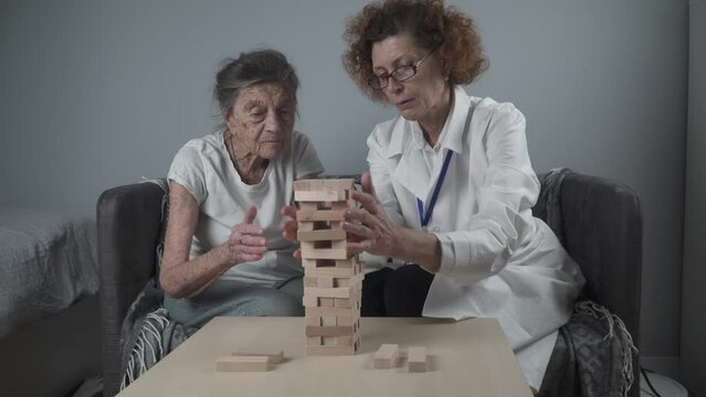 Senior woman playing Jenga, build tower of blocks. Elderly doctor in white lab coat, supporting senior patient, developing logic, fine motor skills, Alzheimer disease at home with old lonely female.