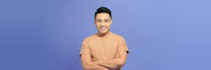 Picture of young happy asian man standing with arms crossed over purple