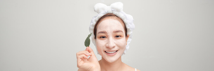 Banner of pretty woman holds green leaf, looks pensively aside, applies clay mask on face,