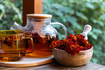 Marigold tea still life on table in green garden background. Healthy hot drink benefits. Natural organic aromatic drink in cup. Home-grown immunity-boosting herbs for tea. Autumn winter warming - obrazy, fototapety, plakaty