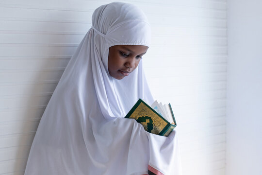 African Muslim child girl wears hijab abaya reading the Quran, leaning against white wall