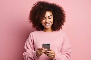 Fototapeta na wymiar Studio portrait of beautiful African American woman with smartphone in pink clothes against pink background. Positive girl with Afro haircut texting message, enjoying online communication, using app.