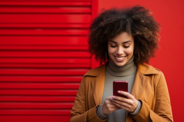 Studio portrait of beautiful African American woman with smartphone in orange clothes against red background. Positive girl with Afro haircut texting message, enjoying online communication, using app.