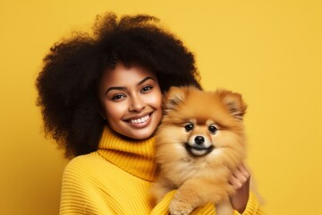 Portrait of smiling young woman with afro hairstyle in yellow sweater posing with cute Pomeranian spitz puppy. Happy girl hugging her beloved pet. Love between human and dog. Yellow studio background. - obrazy, fototapety, plakaty