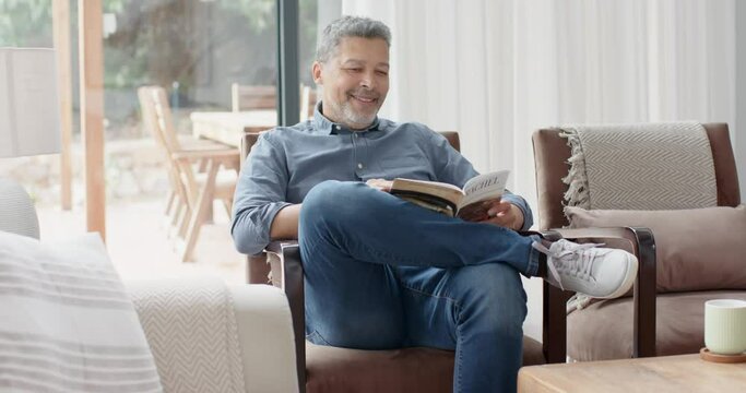 Happy senior biracial man sitting in armchair reading book in living room, slow motion