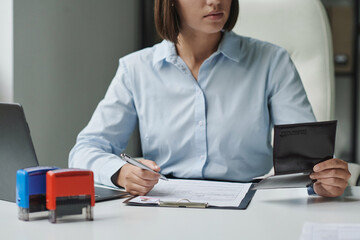 Young specialist checking passport while filling documents for visa at her workplace in office