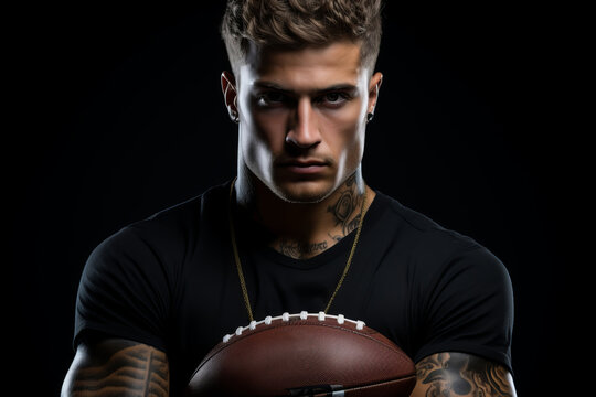 Studio portrait of brutal tattooed professional American football player in black T-shirt holding ovoid ball in his hands. Muscular, confident Caucasian athlete with determined look. Black background.