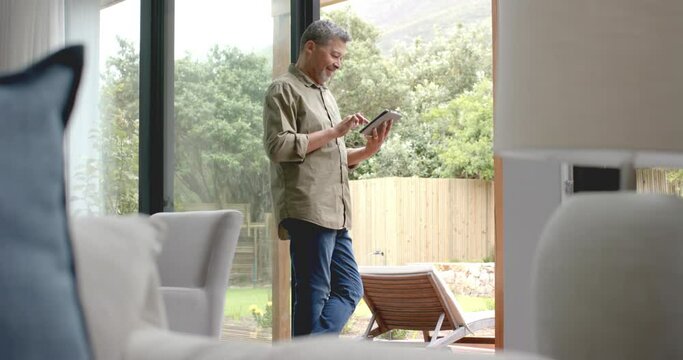 Happy senior biracial man standing by terrace using tablet at home, copy space, slow motion