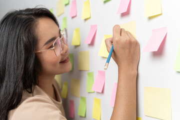 Young smiling business woman, leader, consultant, writing creative work on sticky post it notes on glass wall, female coach, project planning.