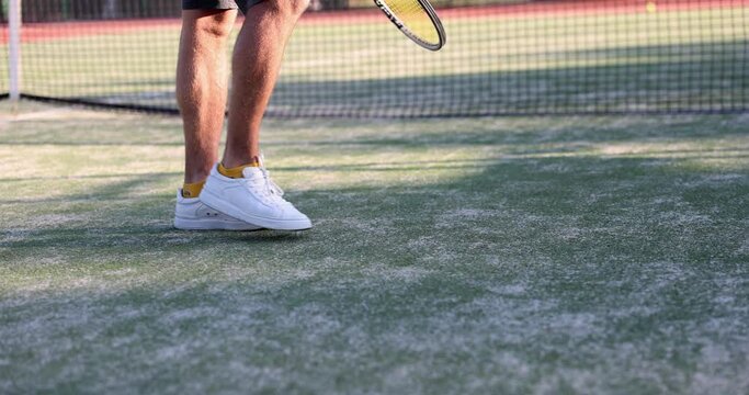 Athlete legs on tennis court holds racket and knocks ball on court. Tennis player walks and hits ball on sports ground