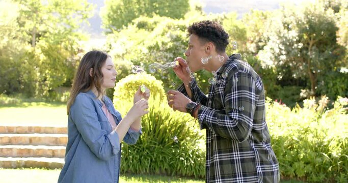 Happy diverse couple blowing bubbles in sunny garden, in slow motion