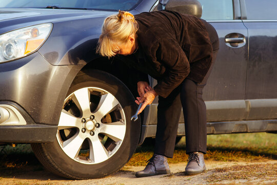 Unrecognizable blonde woman is leaning over car and changing broken tire on autumn day in nature.