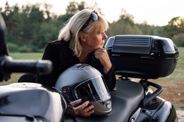 Motorcycle tourism. Mature blonde stands near her motorcycle on autumn day and looks away. Hobbies.