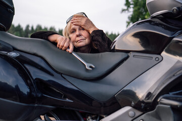 Thoughtfulness and fatigue are shown by mature female with wrench near motorcycle on rural road .