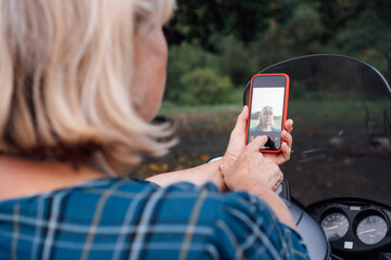 Unrecognizable blonde in blue blouse takes selfie with her phone while sitting on her motorcycle .
