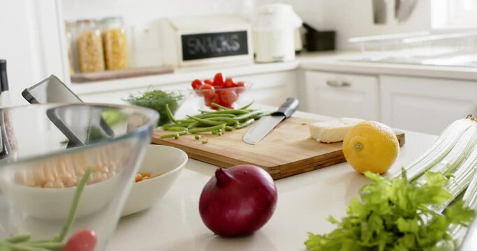 Knife, cutting board, vegetables and tablet on counter in sunny kitchen, slow motion
