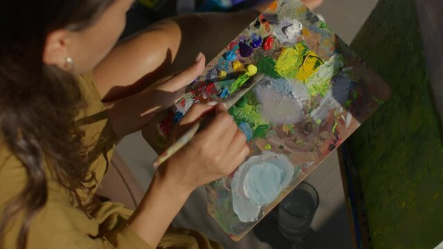A young girl mixes bright acrylic paints on a palette, a brush mixes acrylic paints, a palette is in the hand of a young woman with long brown hair, a shot from above of a girl who paints