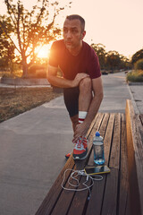 Sporty adult man with ankle pain stretching on a park bench after jogging in sunset time.