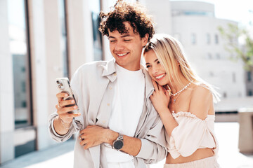 Young smiling beautiful woman and her handsome boyfriend in casual summer clothes. Happy cheerful family. Female having fun. Couple posing in street. Hold smartphone, use phone apps, look at screen