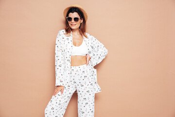 Young beautiful smiling female in trendy summer costume clothes. Carefree woman posing near beige wall in studio. Positive model having fun indoors. Cheerful and happy. In hat and sunglasses