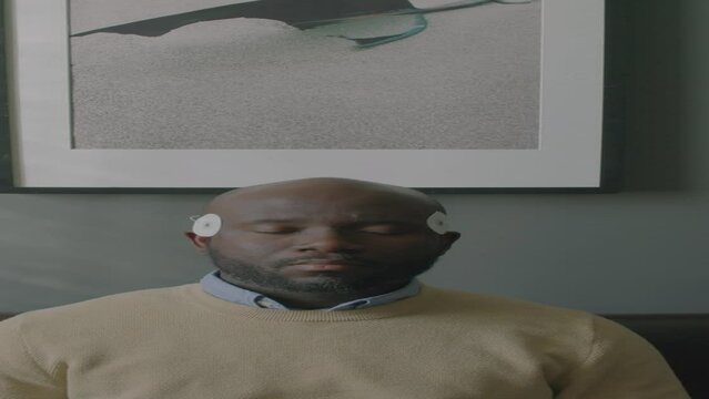 Vertical shot of Black man with electrodes attached to his temples sitting with eyes closed on sofa while getting electroencephalogram during sleep study test in clinic