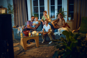 Family watching tv at home. Young woman, man and children sitting on sofa in living room, eating snacks and watching movie in evening. Concept of family, leisure time, relaxation, childhood and