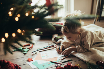Cute girl writing a letter to Santa Claus, lying on a wooden floor under decorated Christmas tree.