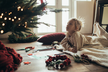 Cute girl writing a letter to Santa Claus, lying on a wooden floor under decorated Christmas tree. - obrazy, fototapety, plakaty