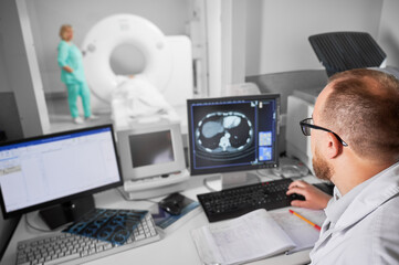 Medical computed tomography or MRI scanner. Back view of male doctor sitting at computer, nurse making MRI on blurred background. Specialists working in clinic. Concept of modern diagnostics. - obrazy, fototapety, plakaty
