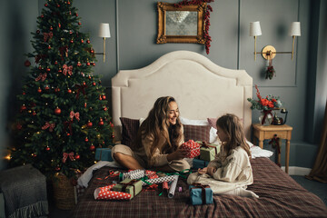 Young beautiful mother and her daughter packing and wrapping presents in a cozy decorated bedroom with Christmas tree. - obrazy, fototapety, plakaty