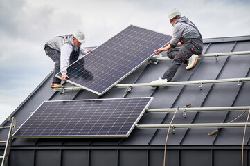 Workers building solar panel system on roof of house. Men technicians in helmets carrying photovoltaic solar module outdoors. Concept of alternative and renewable energy.