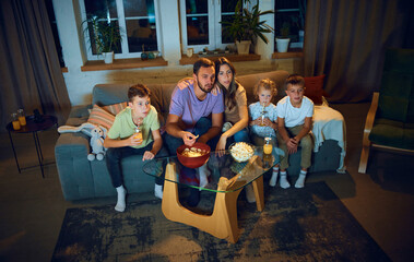 Family watching tv together, parents sand children sitting on couch in living room, eating snacks, watching movie in the evening. Concept of family, leisure time, relaxation, childhood and parenthood - Powered by Adobe