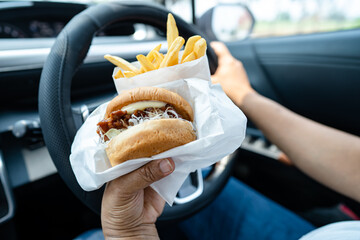 Asian lady holding hamburger and French fries to eat in car, dangerous and risk an accident.