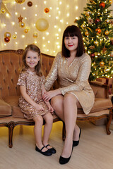 Obraz na płótnie Canvas Happy mother and daughter in golden dresses sitting on sofa in christmas decoration