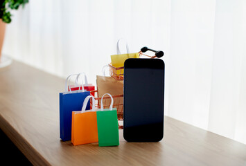 E-commerce and online shopping concept. Shopping cart, mobile phone, colourful bags near the window. Blurred background.
