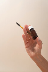 Female hand holding amber glass cosmetic bottle and brow brush on beige background