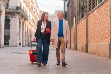 Adult couple enjoying the honeymoon doing a tour in a historical town and smiling. A woman and a men in a weekend trip sightseeing with the suitcase and laughing