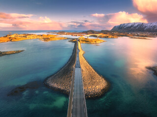 Aerial view of bridge over the sea and snowy mountains in Lofoten Islands, Norway. Fredvang bridges at sunset in winter. Landscape with blue water, rocks in snow, road, sky with clouds. Top drone view - obrazy, fototapety, plakaty